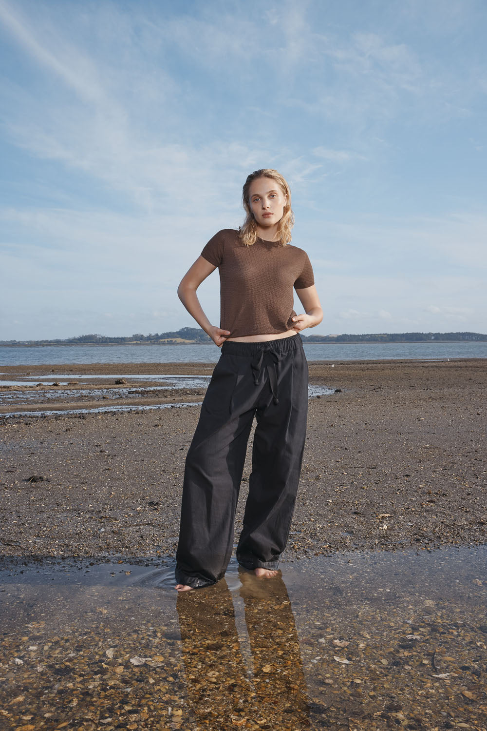 Woman standing on a beach wearing a brown t-shirt and black pants.