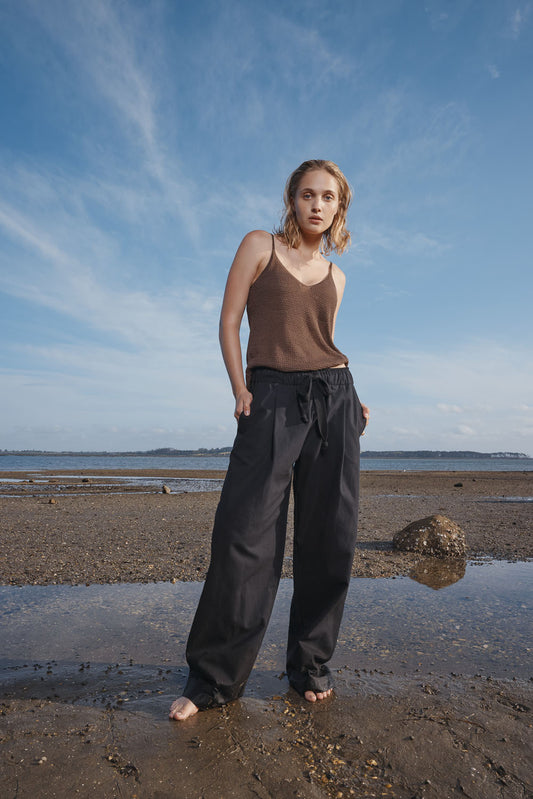 Woman standing on a beach wearing a brown camisole tank top and black pants.