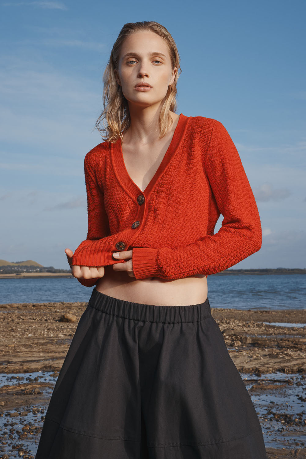 Woman wearing a red cardigan and black skirt standing on a beach with blue sky and water in the background.
