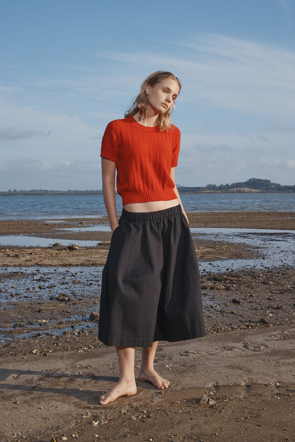 Woman wearing a red knitted top and black skirt standing on a beach with a blue sky.