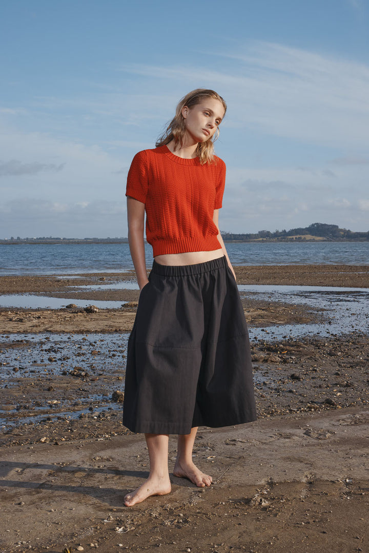 Woman wearing a red knitted top and black skirt standing on a beach with a blue sky.