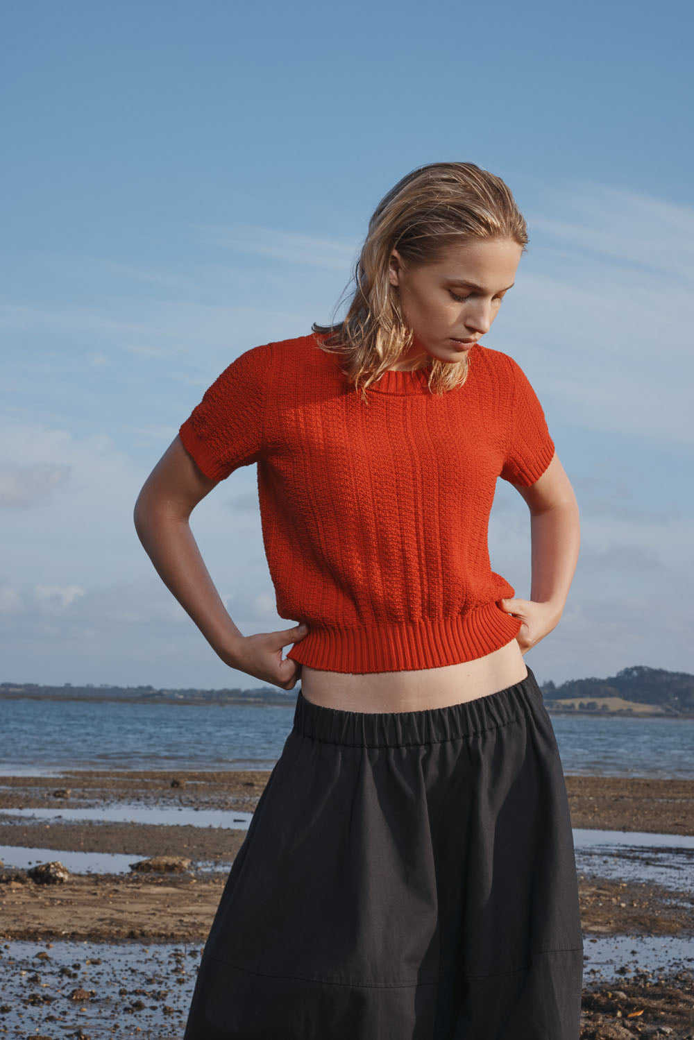 Woman wearing a red knitted top and black skirt standing on a beach.