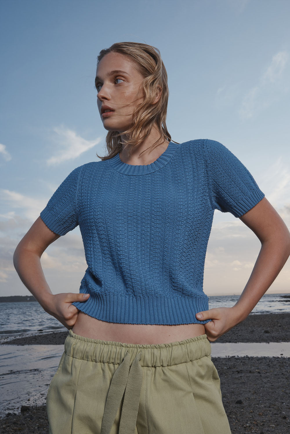 Woman wearing a blue knitted top and light green pants standing on a beach.