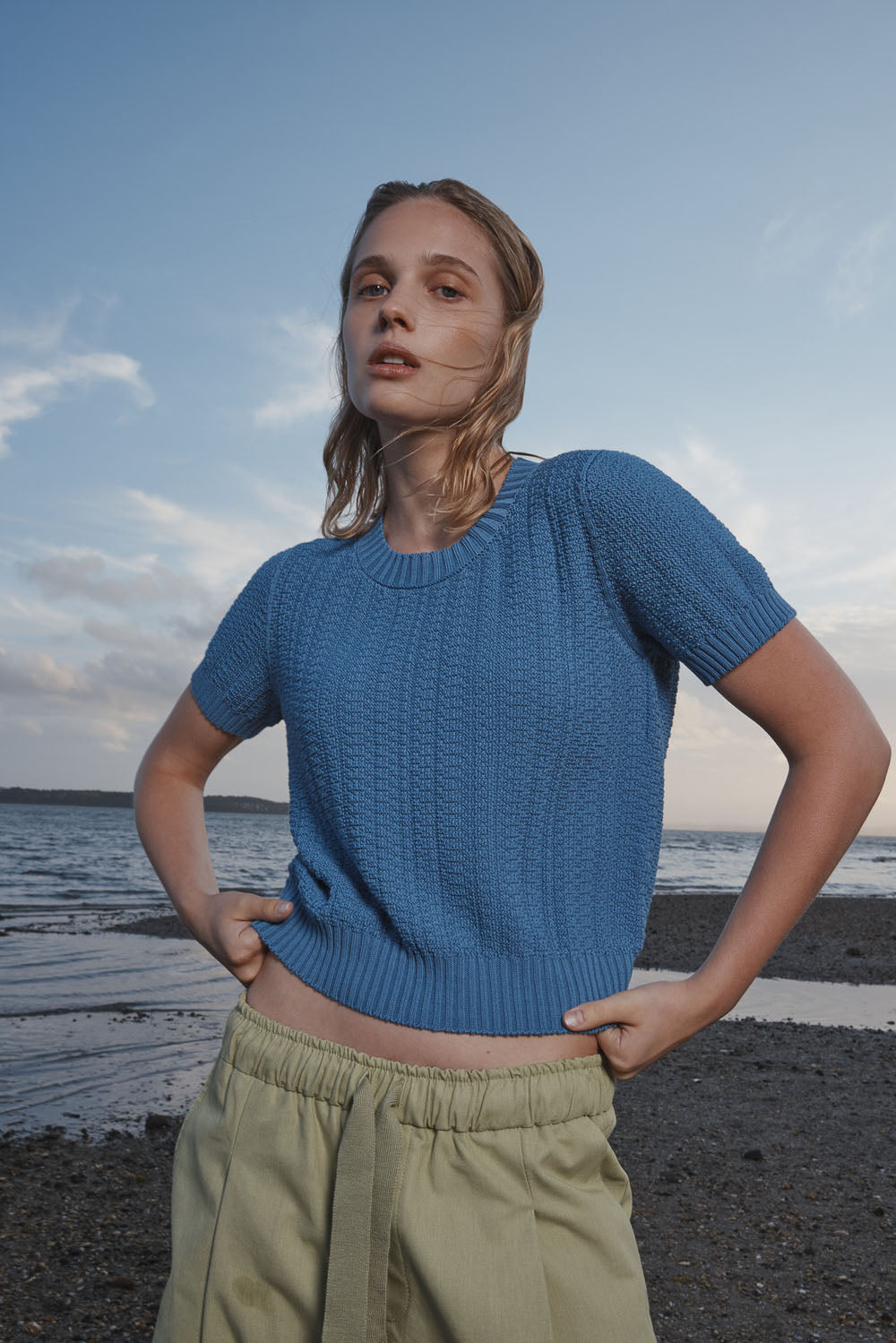 Woman wearing a blue knitted top and light green pants standing on a beach.