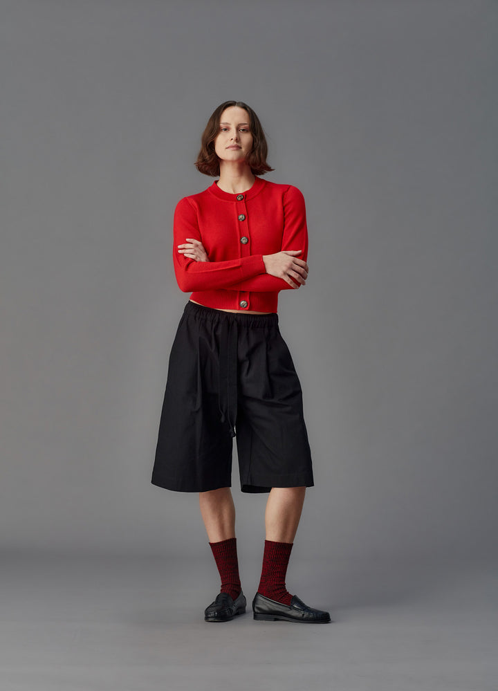 Person wearing a red cardigan, black shorts, and maroon socks on a gray background