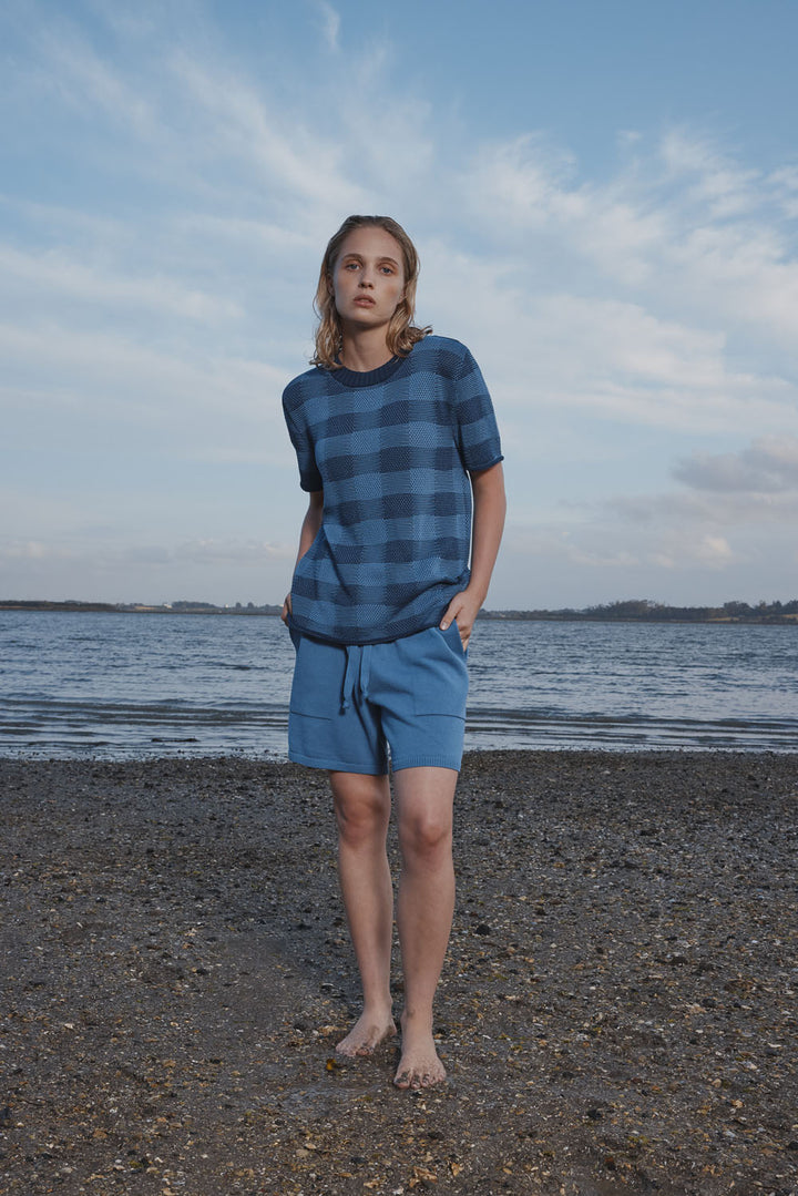 Woman wearing a blue plaid shirt and shorts standing on a beach with water and sky in the background.