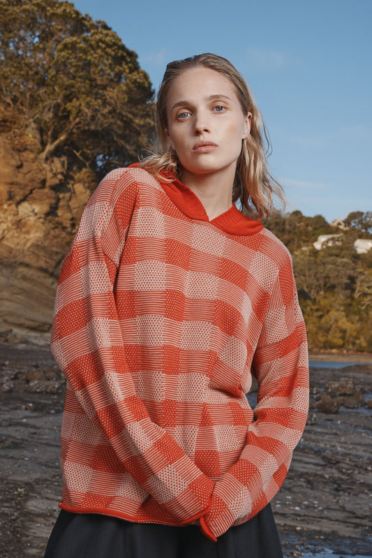 Woman wearing a red and white checkered sweater outdoors with trees and water in the background.