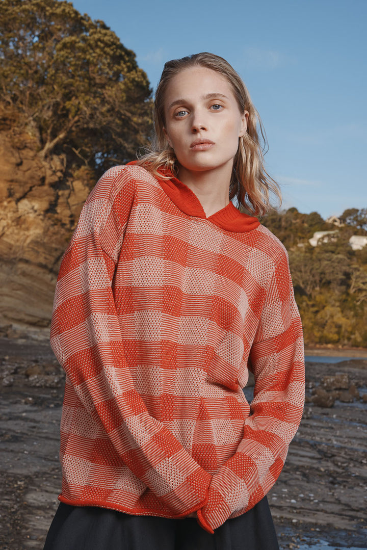 Woman wearing a red and white checkered sweater outdoors with trees and water in the background.