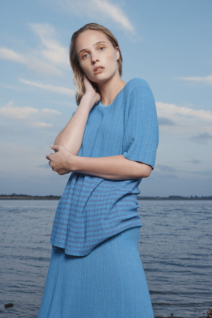 Woman in a knit blue t-shirt and skirt standing by a body of water with a blue sky.