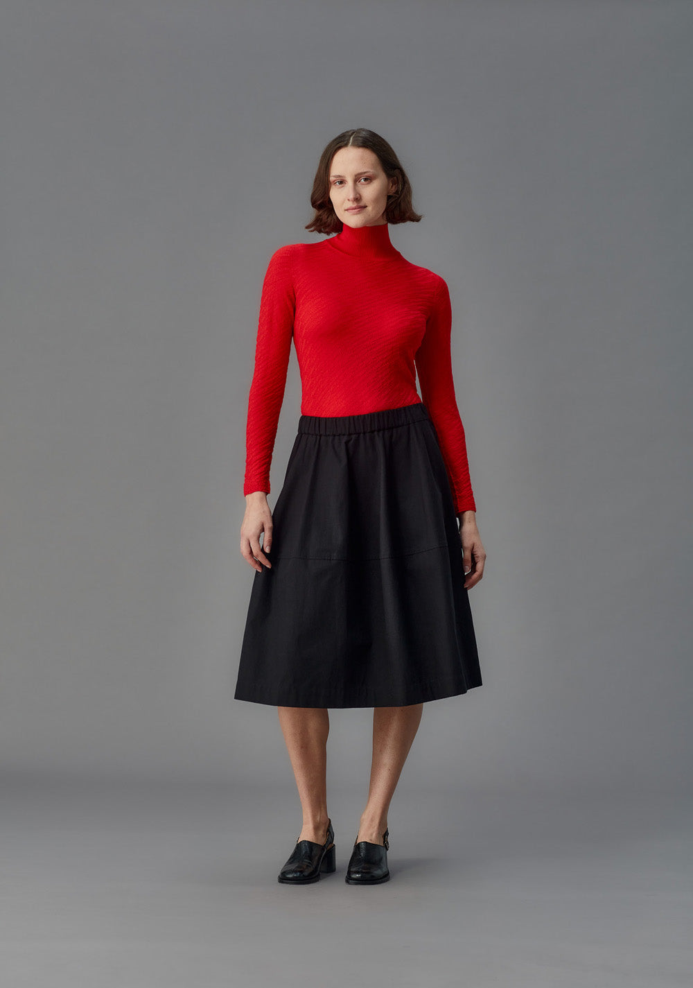 Woman wearing a red turtleneck sweater and black skirt on a gray background