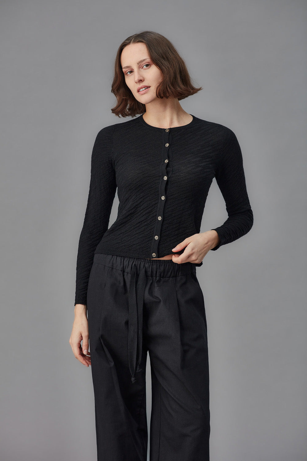 Woman wearing a black cardigan with buttons and black pants on a gray background