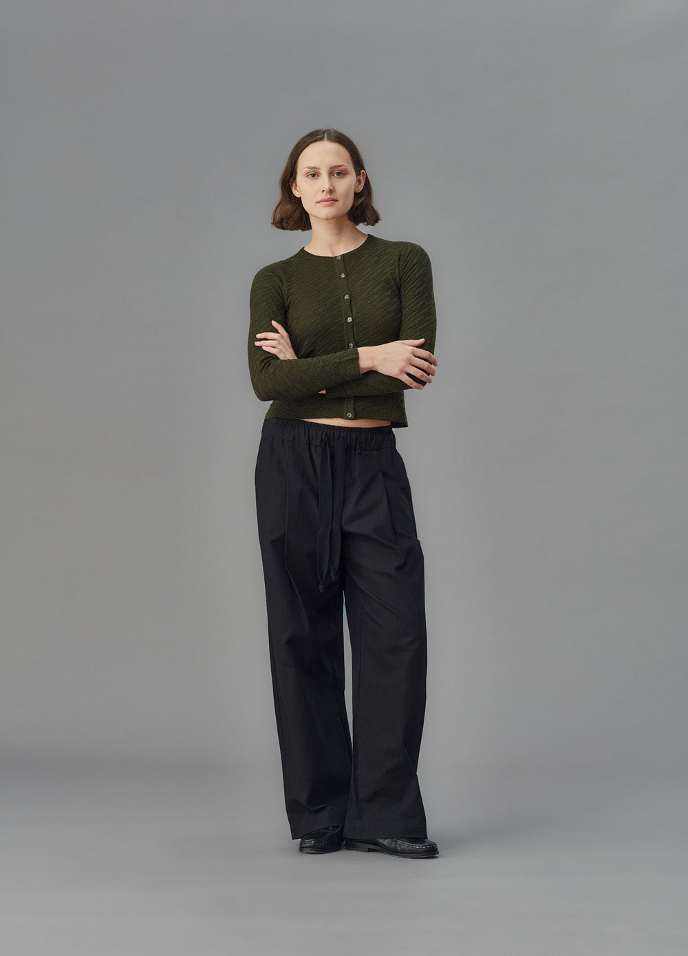 Person wearing a green cardigan and black pants on a gray background