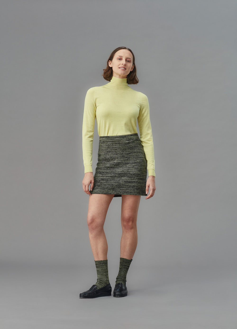 Woman wearing a green knit skirt and light yellow turtleneck sweater against a gray background