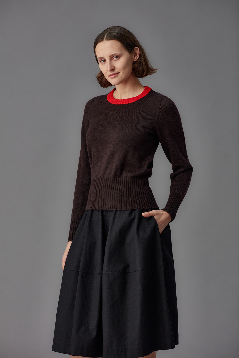 Woman wearing a brown sweater with a red collar and black skirt on a gray background