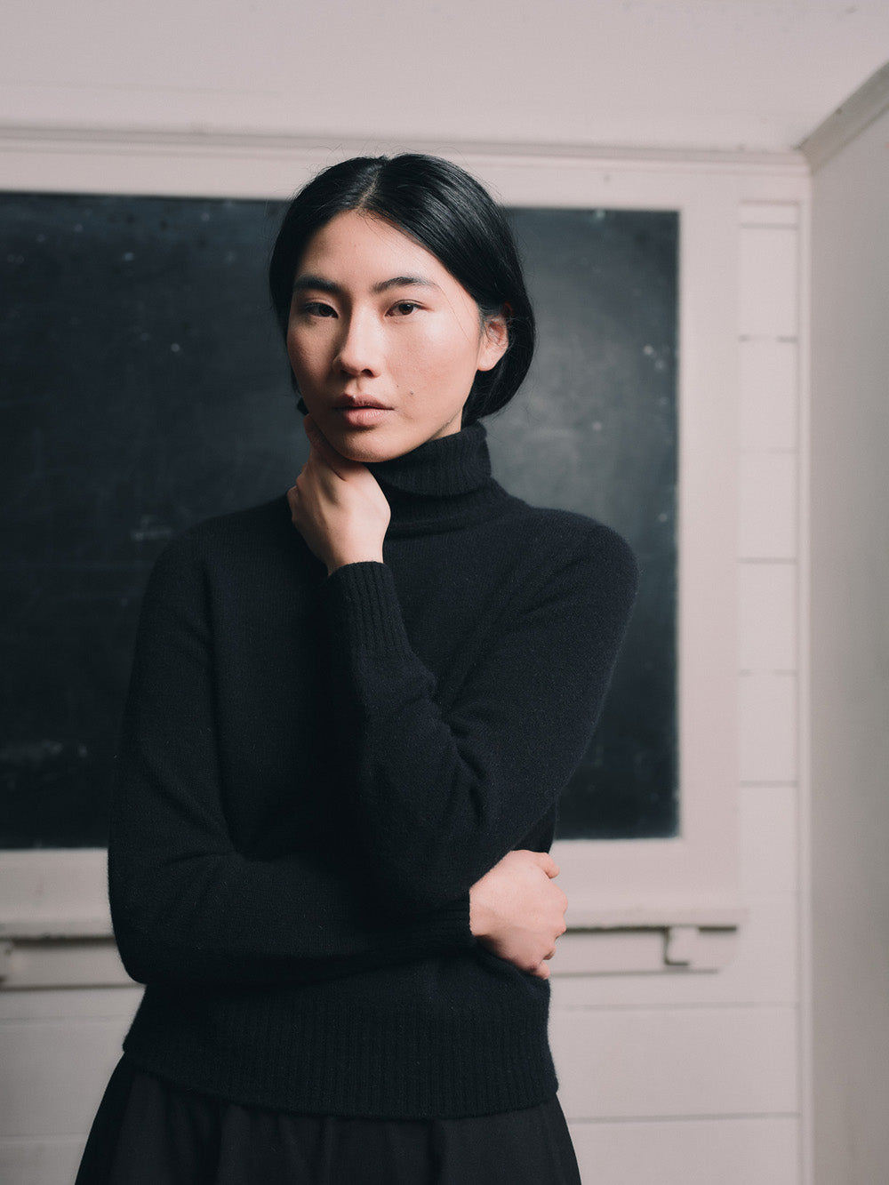 Woman wearing an black funnel neck sweater and black skirt standing in front of a blackboard.