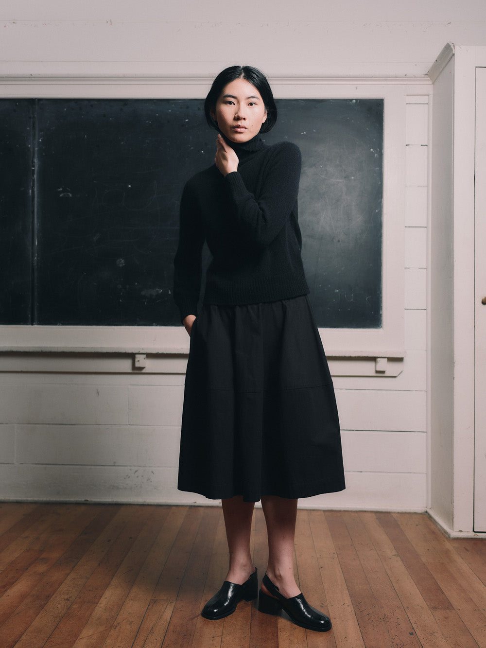 Woman wearing an black funnel neck sweater and black skirt standing in front of a blackboard.