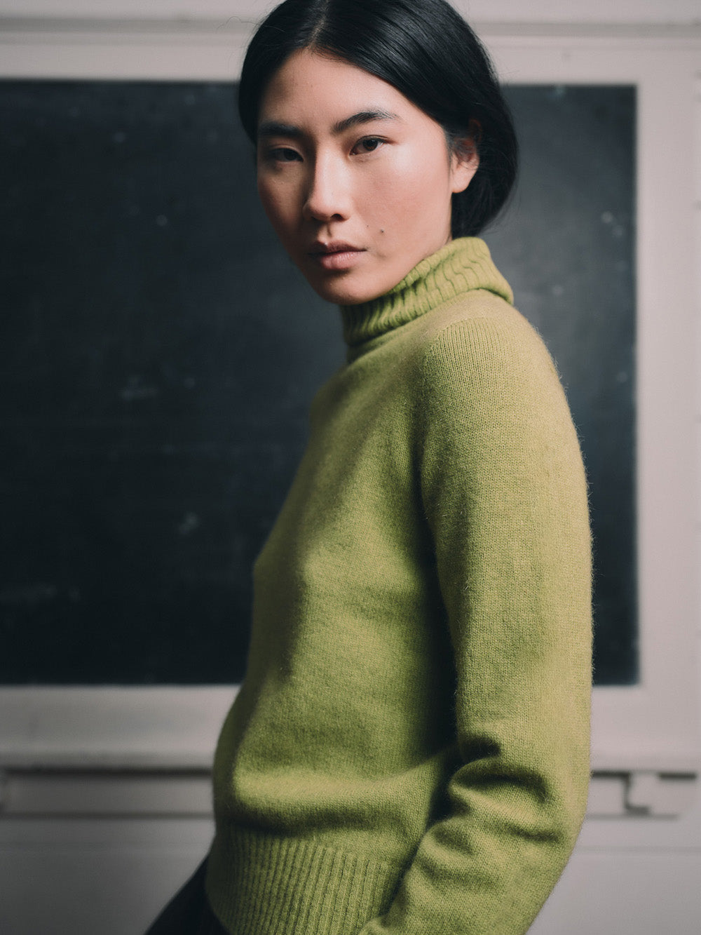 Woman wearing an herb green sweater and brown skirt standing in front of a blackboard.