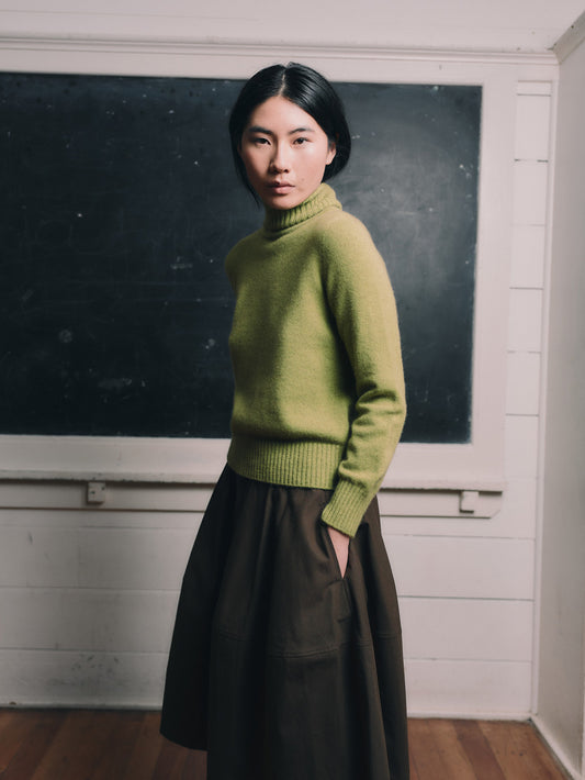 Woman wearing an herb green sweater and brown skirt standing in front of a blackboard.
