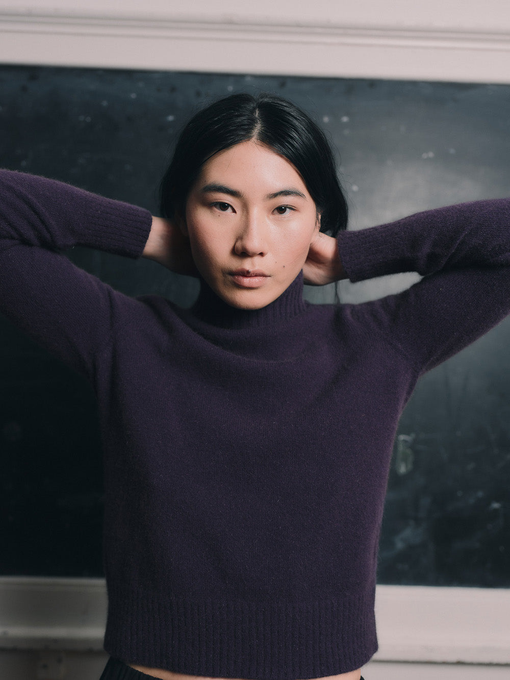 Woman wearing an purple funnel neck sweater and black skirt standing in front of a blackboard.