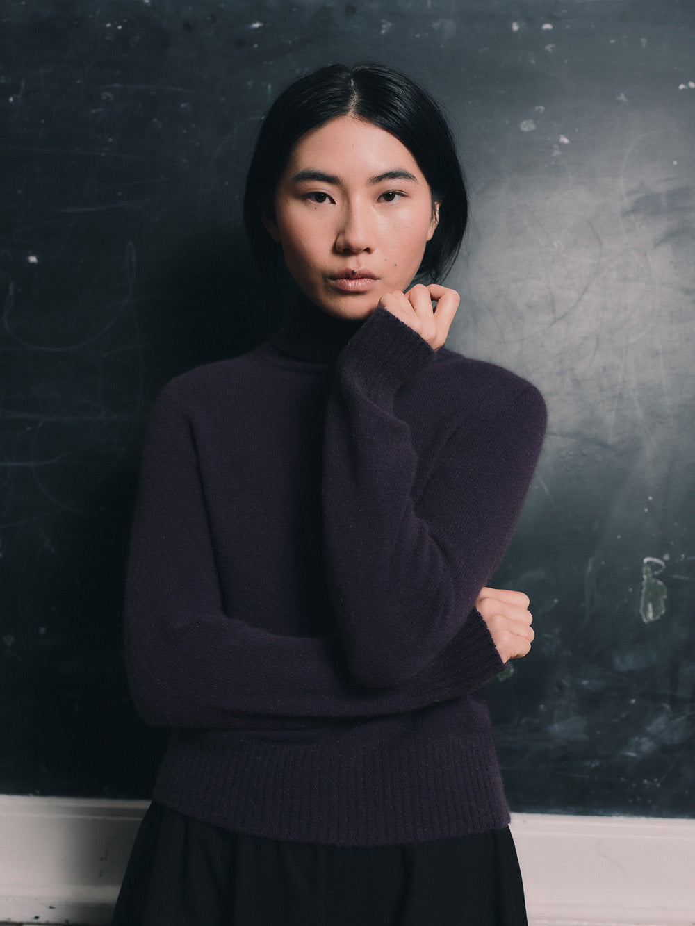 Woman wearing an purple funnel neck sweater and black skirt standing in front of a blackboard.
