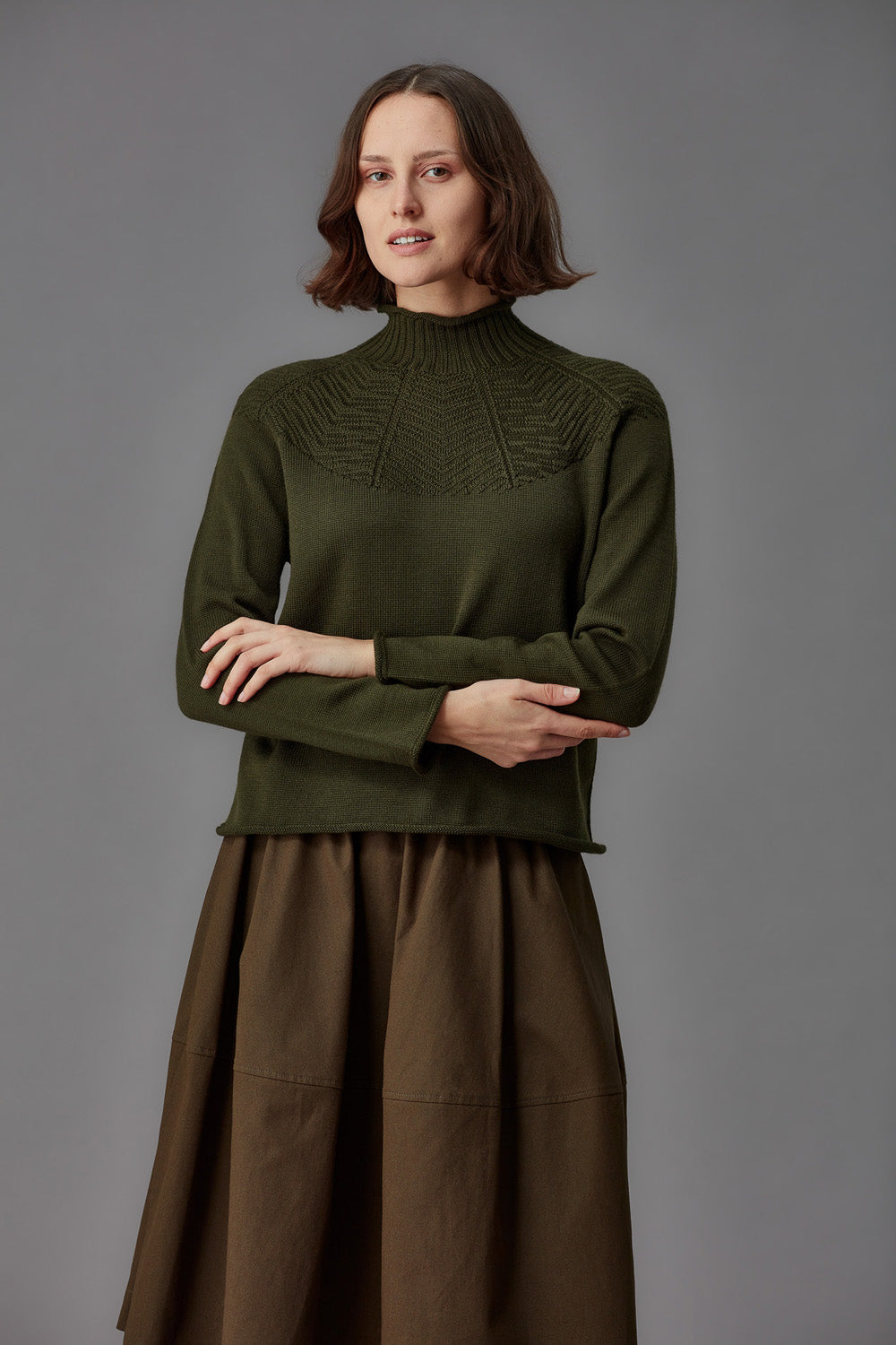 Woman wearing a green mock neck sweater and brown skirt against a gray background