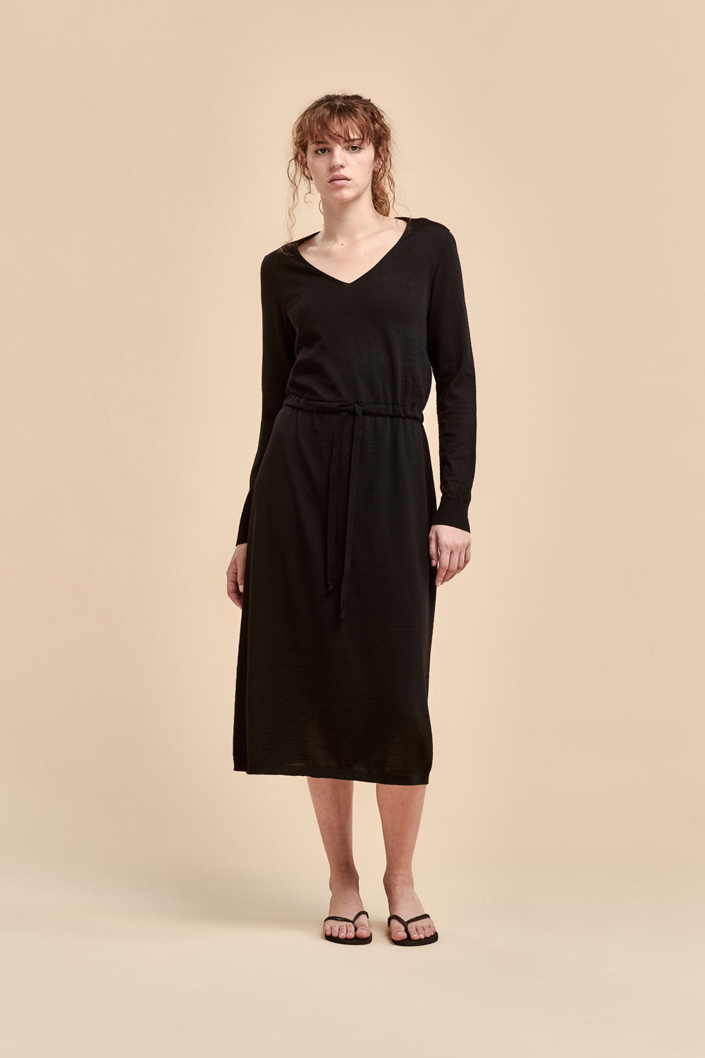 A woman modeling a black long-sleeve merino dress with a V-neck and a drawstring belt at the waist, set against a solid beige studio background.