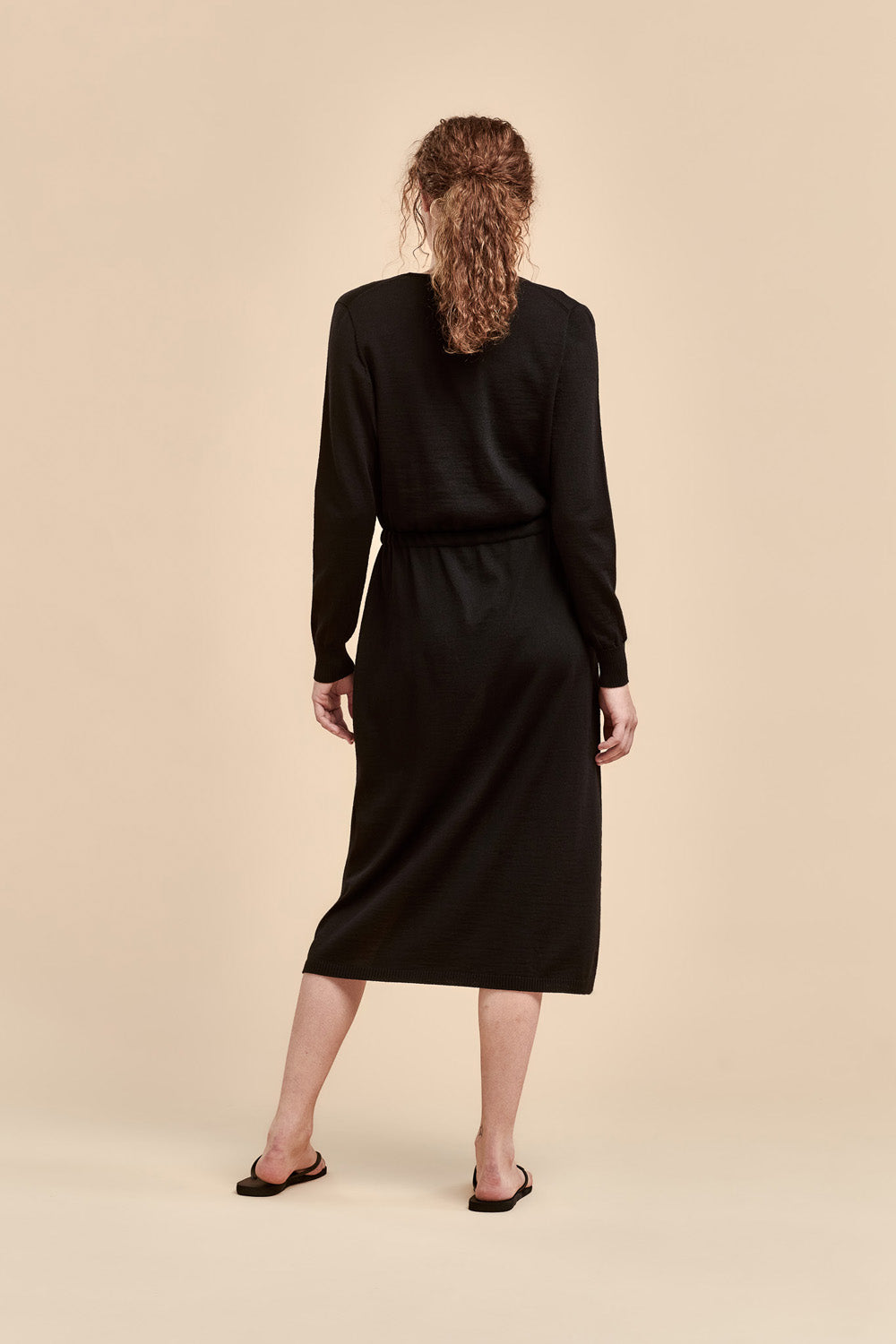 A woman viewed from behind wearing a black long-sleeve merino dress with a V-neck and a drawstring belt at the waist, set against a solid beige studio background.