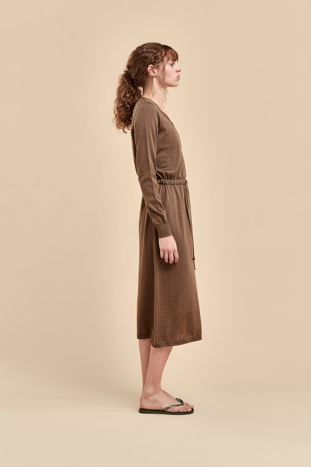 A woman stands in profile wearing a brown long-sleeve merino dress with a V-neck and a drawstring tie, standing against a solid beige studio background.