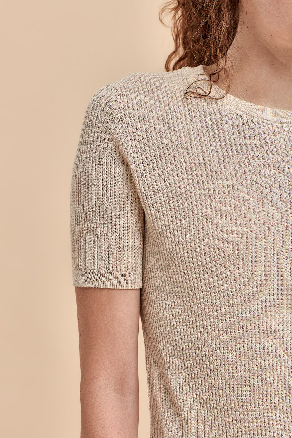 Close-up of a woman wearing a short-sleeve ribbed knit top in a light beige color against a solid beige background.