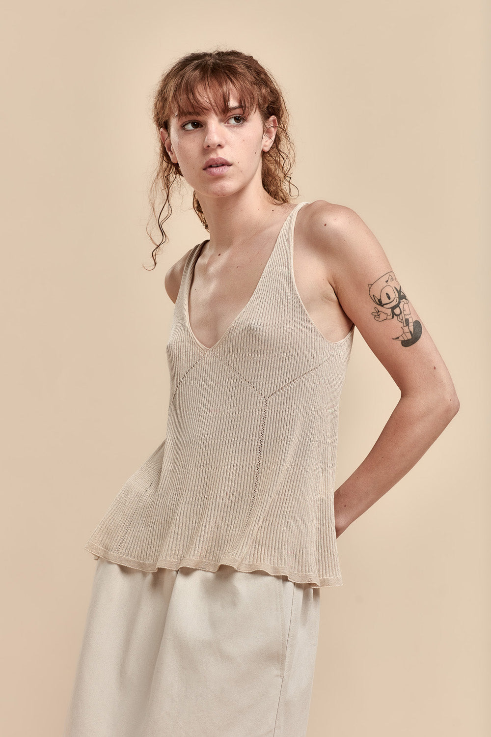 A woman modeling a beige ribbed knit camisole top with a deep V-neck, paired with matching beige trousers, against a solid beige background.