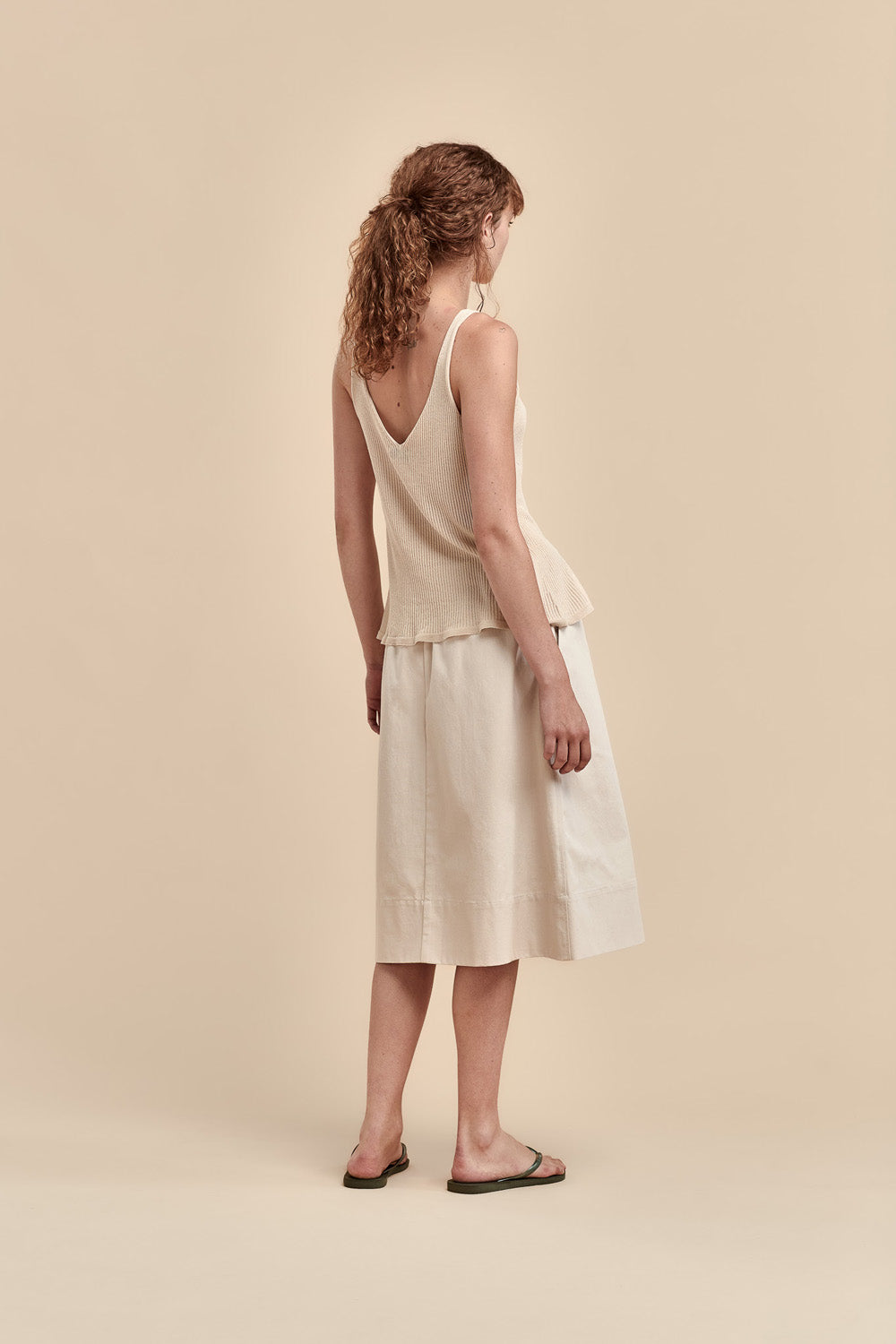 A woman stands with her back to the camera wearing a light beige sleeveless top and matching midi skirt, paired with dark sandals, against a solid beige studio background.