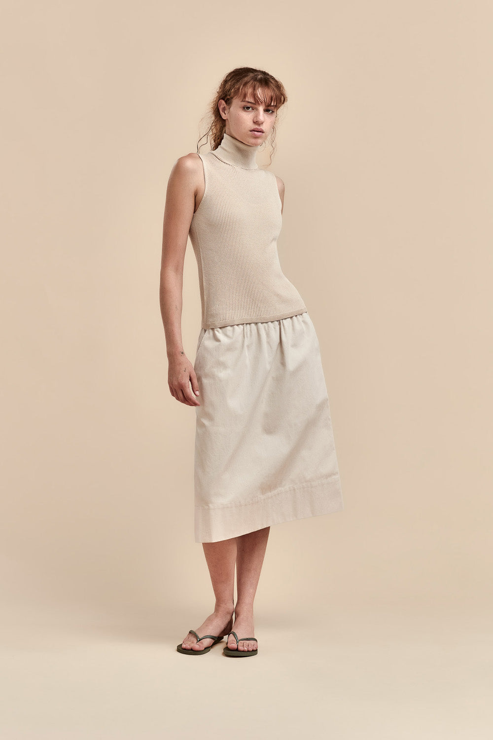 A model stands against a solid beige studio background wearing a sleeveless cream turtleneck top and a matching cream midi skirt with dark brown sandals.
