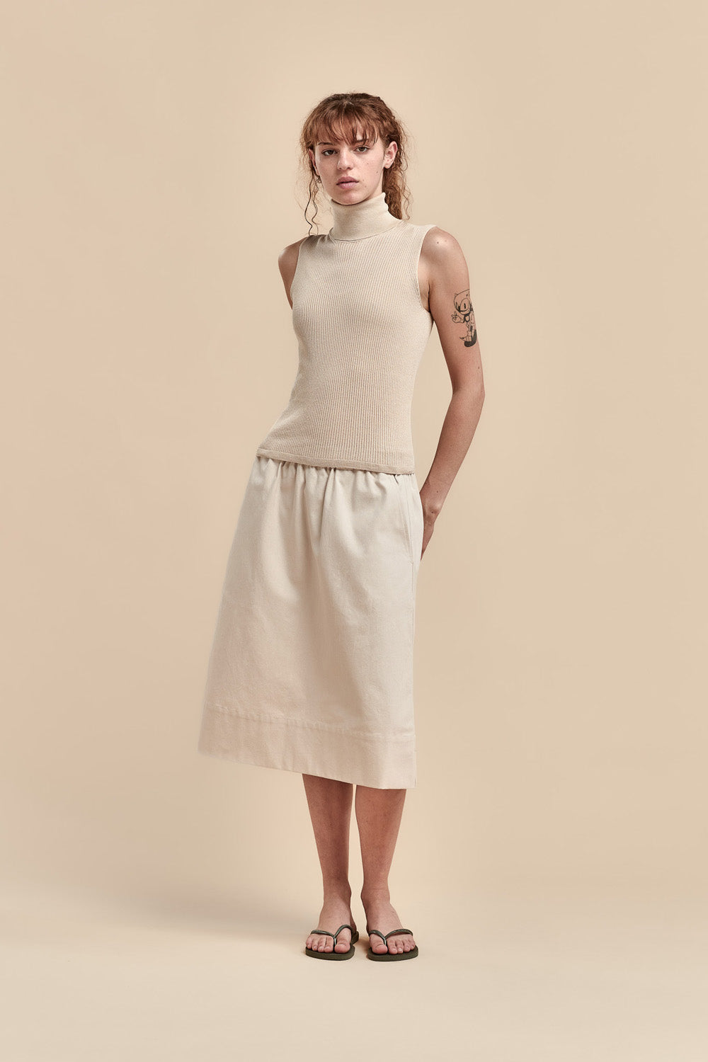 A model stands against a solid beige studio background wearing a sleeveless beige mock-neck knit top and a matching beige midi skirt, paired with dark sandals.
