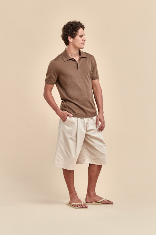 A man wearing a brown merino short-sleeve polo shirt and off-white shorts, standing in profile against a solid beige background.
