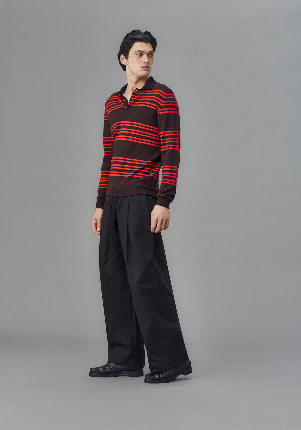Man wearing a brown and red striped sweater with black pants on a gray background