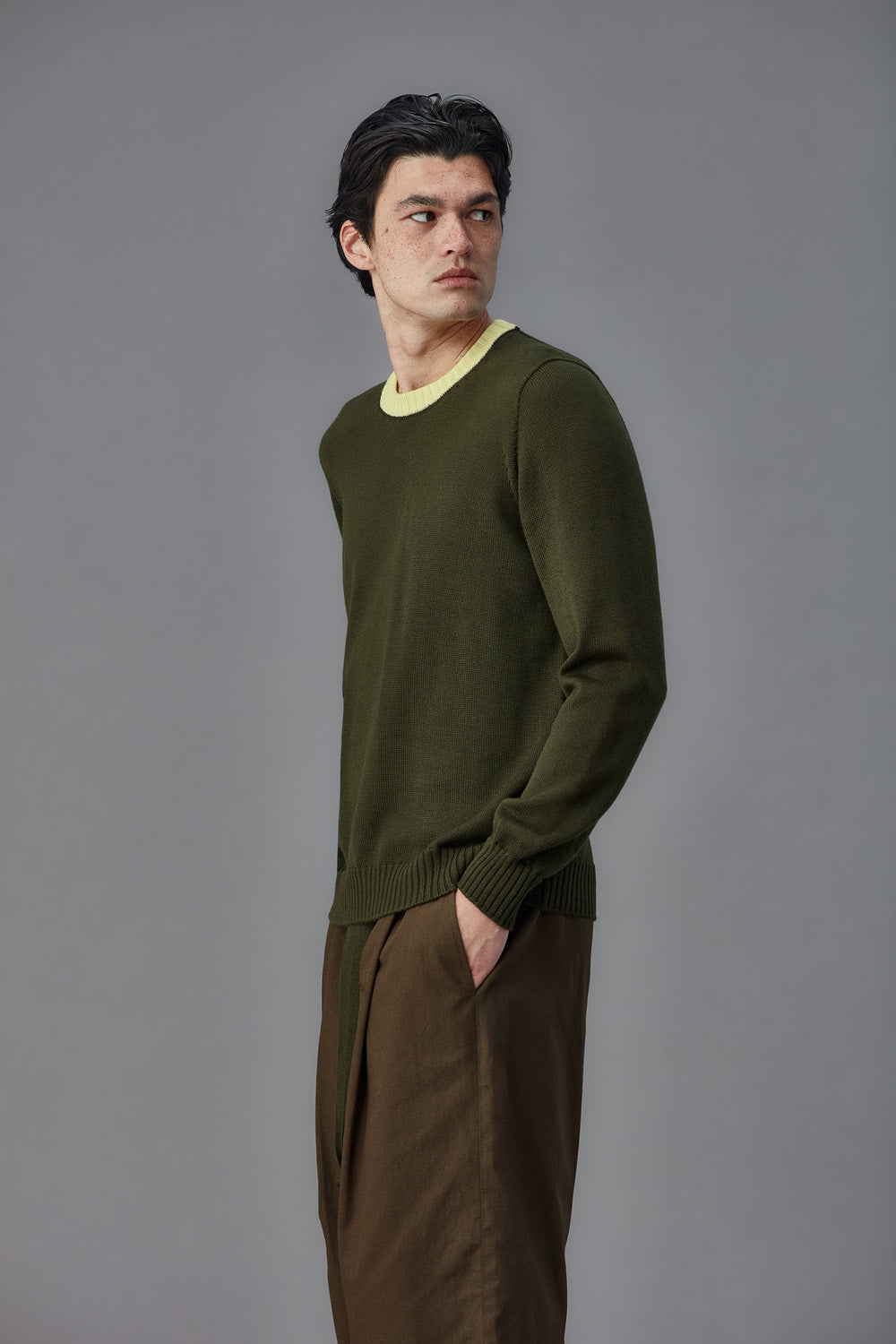Man wearing a green sweater with contrast yellow collar and brown pants against a gray background