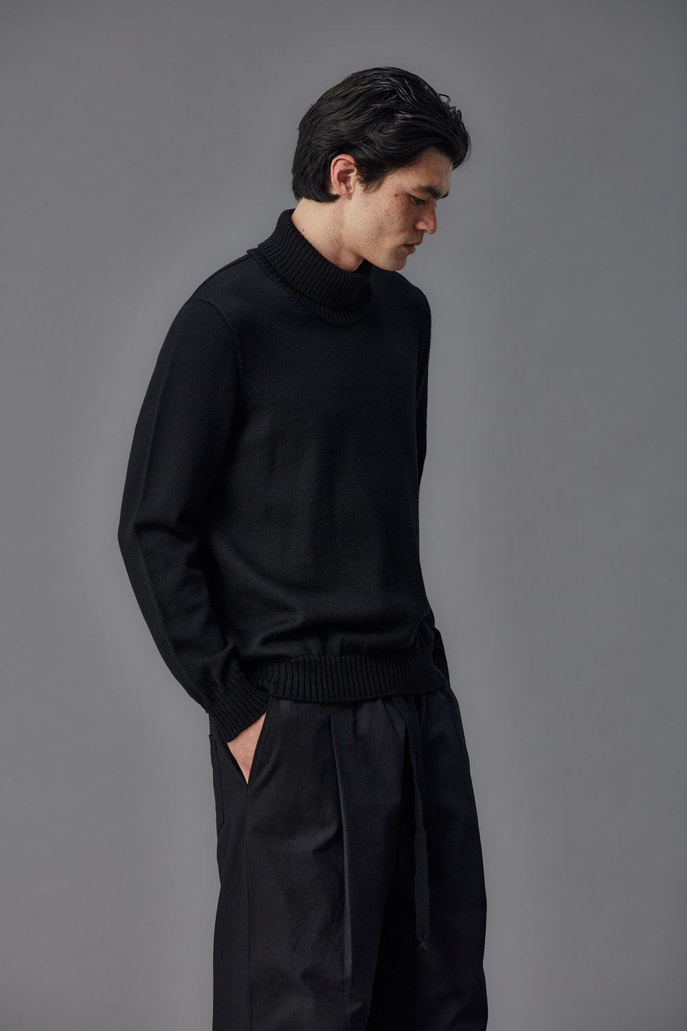 Man wearing a black turtleneck sweater and dark pants against a gray background