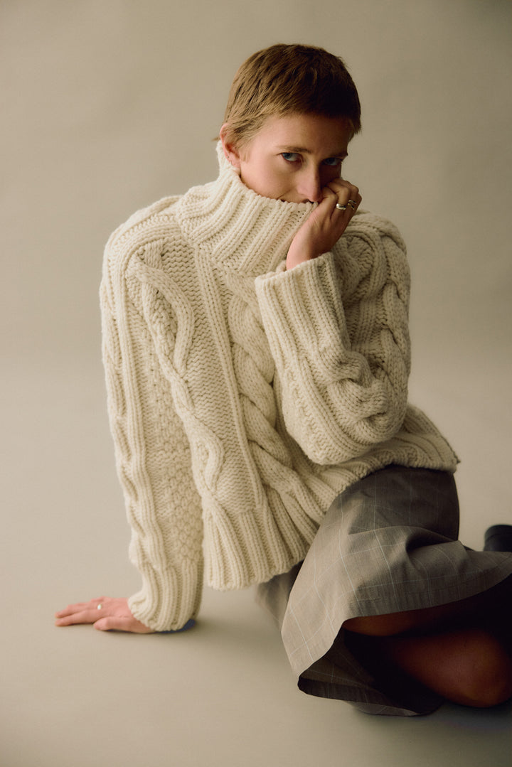 Woman wearing a large cream-coloured cable knitted sweater on a plain background
