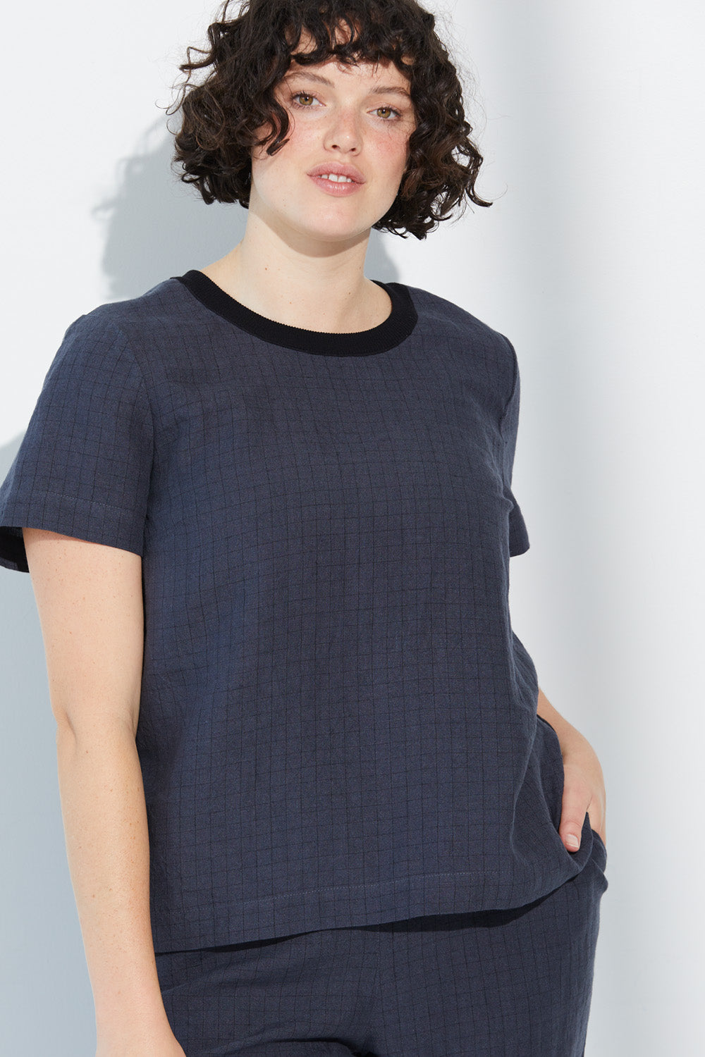 A woman with hand in pocket wearing a charcoal-coloured Standard Issue Grid Linen Tee.